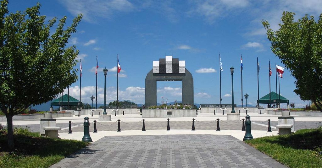 Statues of soldiers storming the beach at the National D-Day Memorial in Bedford, Virginia, with the Overlord Arch towering in the background under a clear blue sky.