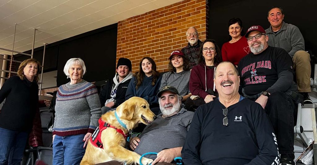 Group of eleven people and one golden retriever service dog smiling together in a casual indoor setting.